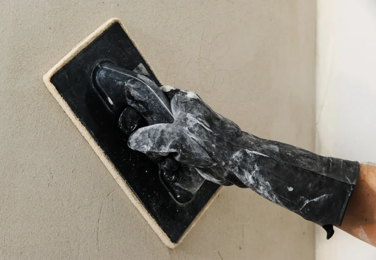 Hand mit schwarzem Arbeitshandschuh führt ein Filzbrett über feuchten Putz an einer Wand.
