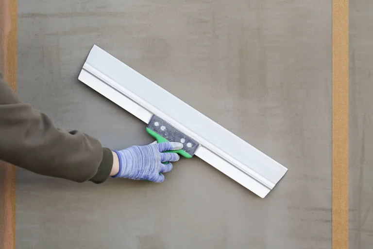 Hand mit blauem Schutzhandschuh glättet mit einer breiten Flächenspachtel feuchten Glattputz an einer Wand.
