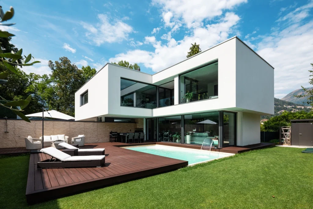 Moderne, weiße Villa mit Glattputzfassade, großen Glasfronten und Pool auf einer dunklen Holzterrasse im Garten.