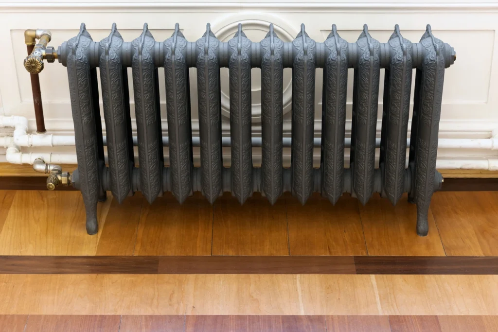 Freistehender Gliederheizkörper aus dunklem Gusseisen mit Ornament-Verzierungen auf Holzparkett vor einer weißen Wandvertäfelung.