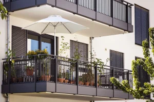 Moderner Hängebalkon mit anthrazitfarbenem Geländer, Sonnenschirm und Topfpflanzen an einer weißen Hausfassade.