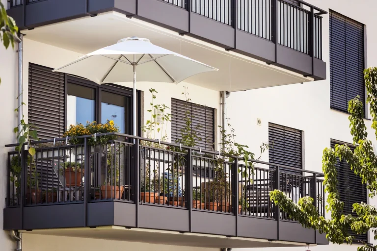 Moderner Hängebalkon mit anthrazitfarbenem Geländer, Sonnenschirm und Topfpflanzen an einer weißen Hausfassade.