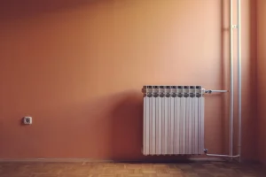 Weißer Lamellenheizkörper an einer orangefarbenen Wand mit Parkettboden, seitlichen Rohren und einer Steckdose links.