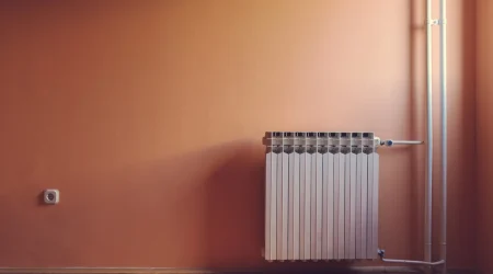 Weißer Lamellenheizkörper an einer orangefarbenen Wand mit Parkettboden, seitlichen Rohren und einer Steckdose links.