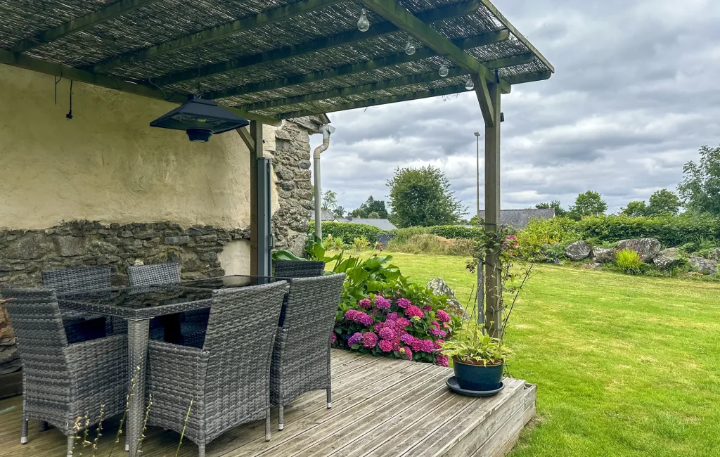 Rustikale Holzterrasse mit einer gemütlichen Sitzgruppe aus grauen Polyrattan-Sesseln unter einer hölzernen Pergola mit Reetdach; im Hintergrund erstreckt sich eine weitläufige grüne Rasenfläche mit blühenden Hortensien.
