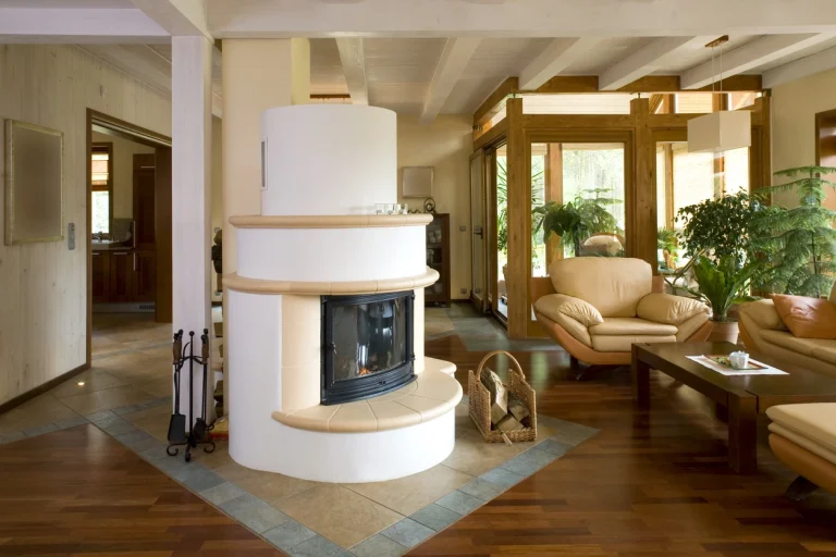 Weißer, runder Kachelofen mit Glaseinsatz in einem Wohnzimmer mit Parkettboden, Ledersesseln und Holzbalkendecke.