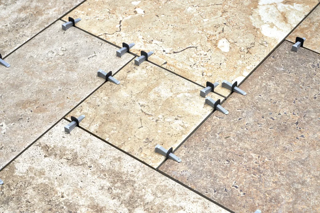 Nahaufnahme der Verlegung von großformatigen Travertin-Fliesen im Modulverband. In den Fliesenfugen sind graue Nivelliersystem-Abstandshalter und Keile eingesetzt, um eine gleichmäßige Fugenbreite und plane Oberfläche des Natursteinbodens zu gewährleisten.