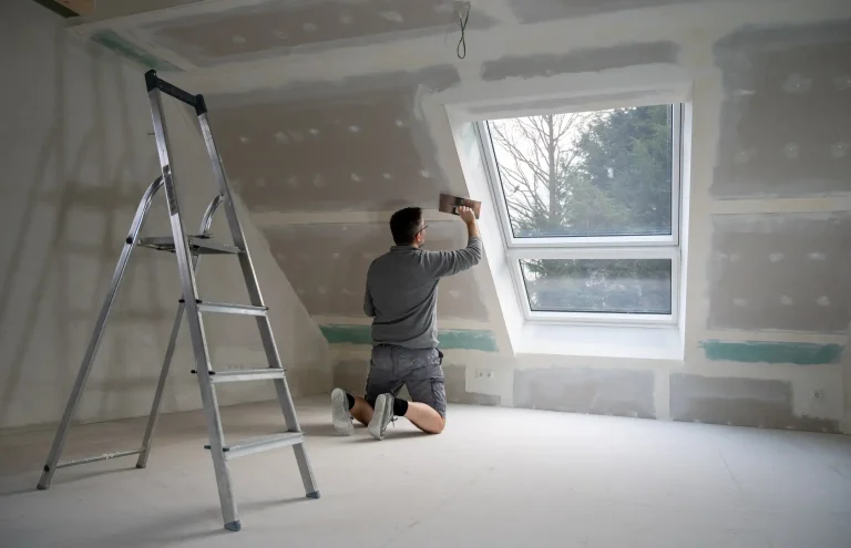 Ein Handwerker kniet im Dachgeschoss und nutzt eine Glättkelle, um die Fugen von Rigips-Platten an einer Dachschräge neben einem Dachfenster zu verputzen. Im Raum steht eine silberne Aluminium-Stehleiter.