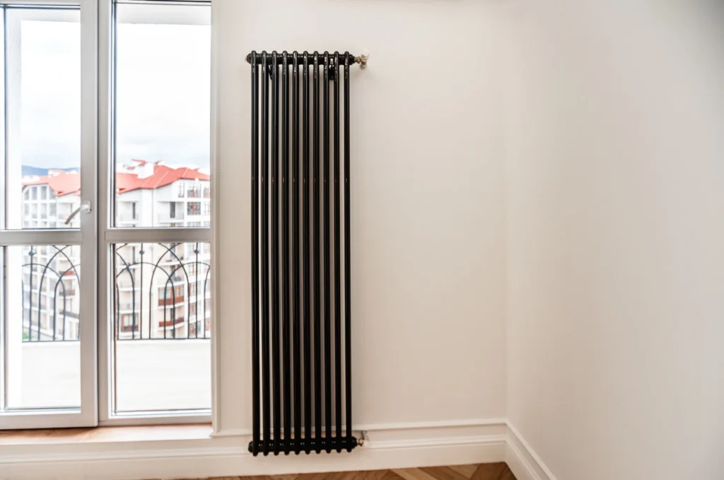 Hoher, vertikaler Röhrenheizkörper in Schwarz an einer weißen Wand neben einer Balkontür mit Blick auf Stadtgebäude.