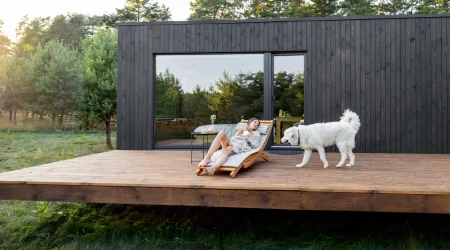 Frau auf Holzliege und weißer Hund auf einer erhöhten Holzterrasse vor einem schwarzen modernen Modulhaus im Grünen.