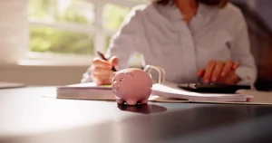 Eine Frau sichtet ihre Finanzunterlagen. Auf den Dokumenten liegt ein Taschenrechner. Vor dem Ordner steht ein Sparschwein.