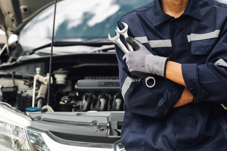 Ein Mechaniker lehnt an einer geöffneten Motorhaube und hält zwei Schraubenschlüssel in der Hand.