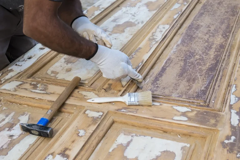 Nahaufnahme eines Handwerkers in weißen Schutzhandschuhen, der mit einem Spachtel alte Farbreste von einer massiven Holztür entfernt. Auf der Tür liegen ein Hammer und ein Pinsel als Werkzeuge für die Untergrundvorbereitung.
