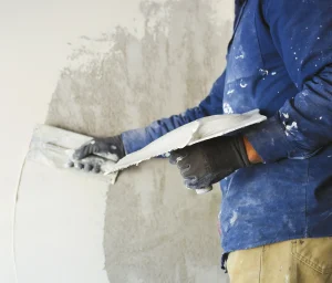 Ein Handwerker in blauer Arbeitskleidung trägt mit einer Glättkelle frischen grauen Putz auf eine Innenwand auf. In der anderen Hand hält er ein Reibebrett mit Putzmörtel zur Vorbereitung der Wandoberfläche.