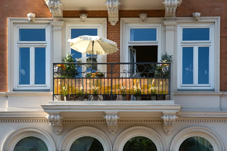 Ein klassischer Vorstellbalkon an einer historischen Altbau-Ziegelfassade, gestützt durch dekorative Konsolen. Die Balkongestaltung umfasst ein schwarzes Schmiedeeisengeländer mit gelbem Sichtschutz, einen hellen Sonnenschirm und diverse Grünpflanzen.