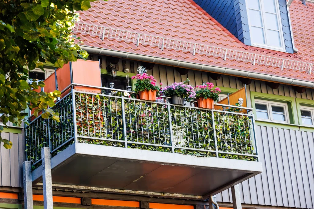 Ein moderner Vorstellbalkon aus Metall, der als Grünoase gestaltet ist. Das Geländer ist mit einer dichten künstlichen Efeu-Hecke als Sichtschutz verkleidet und mit bepflanzten Blumenkästen voller rosa Geranien geschmückt.
