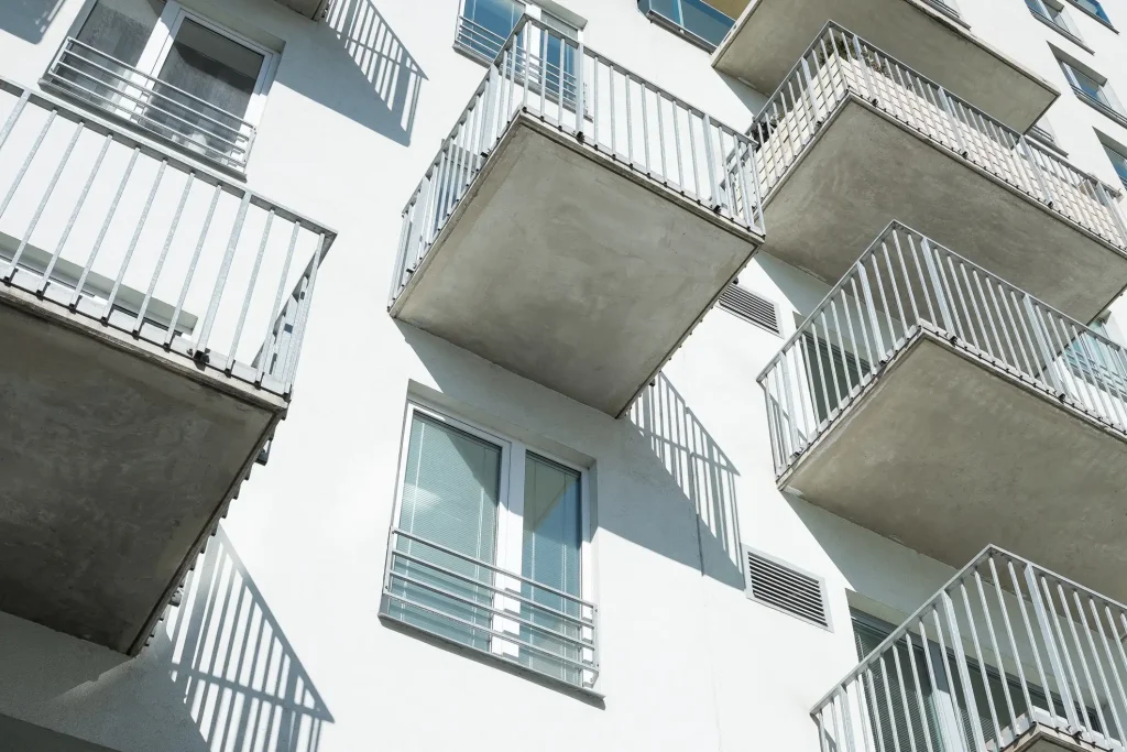 Mehrere schlichte Vorstellbalkone aus Beton und verzinktem Stahl an einer weißen Wohnhaus-Fassade. Die Untersicht verdeutlicht die Konstruktion der Balkonplatten und die einfachen Metallgeländer aus vertikalen Stäben.
