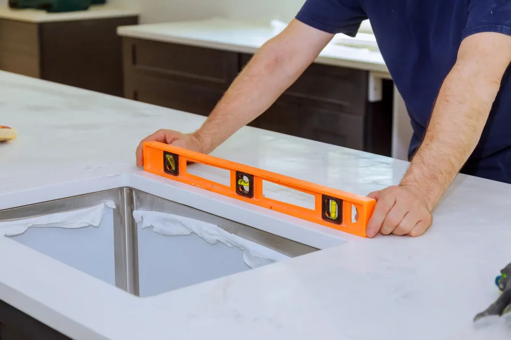 Ein Handwerker überprüft bei der Küchenmontage mit einer orangefarbenen Wasserwaage die waagerechte Ausrichtung einer hellen Küchenarbeitsplatte aus Stein oder Quarzkomposit direkt neben dem Ausschnitt für die Unterbauspüle.