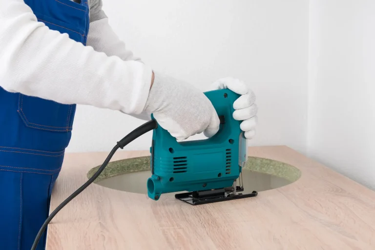 Ein Handwerker in blauem Blaumann und weißen Arbeitshandschuhen sägt mit einer türkisfarbenen Stichsäge einen runden Ausschnitt für ein Spülbecken oder Kochfeld in eine hölzerne Küchenarbeitsplatte.