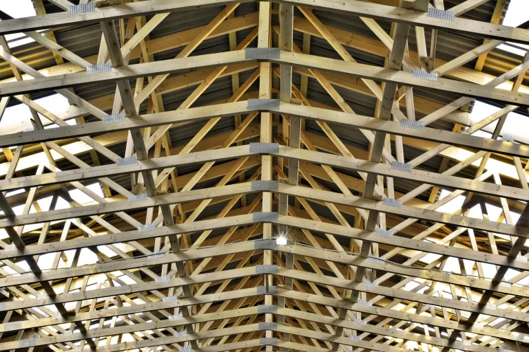 Blick von unten in ein freitragendes Binderdach einer großen Halle. Die symmetrische Holzkonstruktion zeigt aneinandergereihte Nagelplattenbinder aus hellen Holzbalken, die durch metallene Nagelplatten fest miteinander verbunden sind.