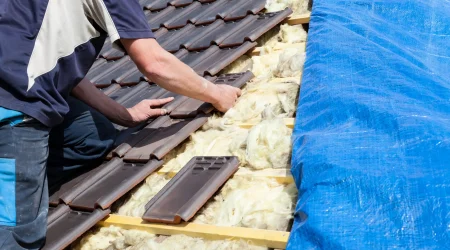 Dachdecker bei der Dacheindeckung während einer Dachsanierung: Ein Handwerker verlegt dunkle Dachziegel auf der hölzernen Lattung. Daneben ist die gelbe Dachdämmung aus Mineralwolle zwischen den Sparren sichtbar, während die rechte Dachseite mit einer blauen Abdeckplane geschützt ist.