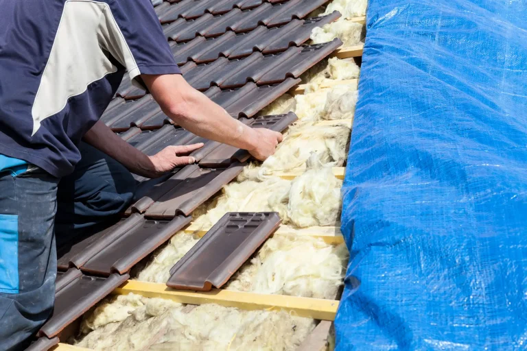 Dachdecker bei der Dacheindeckung während einer Dachsanierung: Ein Handwerker verlegt dunkle Dachziegel auf der hölzernen Lattung. Daneben ist die gelbe Dachdämmung aus Mineralwolle zwischen den Sparren sichtbar, während die rechte Dachseite mit einer blauen Abdeckplane geschützt ist.