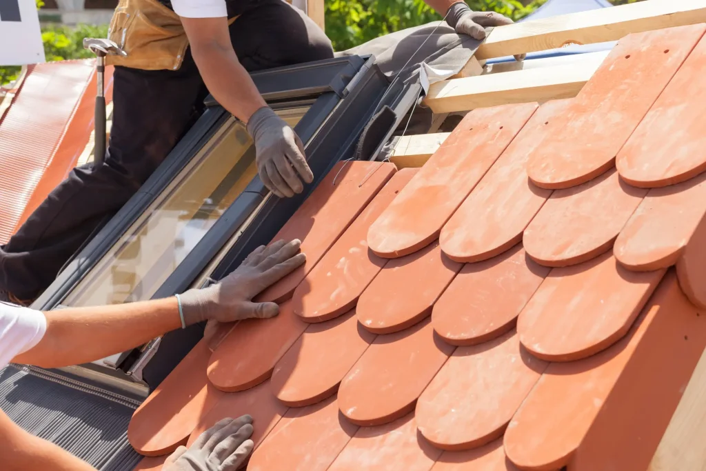 Dachdecker arbeiten an einer Dacheindeckung mit Biberschwanz-Dachziegeln direkt neben einem neu eingebauten Dachfenster. Zwei Dachdecker richten die roten, abgerundeten Tondachziegel in Kronendeckung auf der hölzernen Traglatte aus.