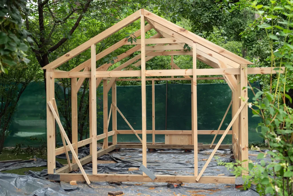 Die Außenansicht zeigt das rohe Holzskelett eines freistehenden Gartenhauses im Bau. Der Rahmen besteht aus unbehandeltem Bauholz mit einem Satteldach-Giebel und steht auf einer schwarzen Bodenschutzfolie inmitten üppiger, grüner Gartenbepflanzung.
