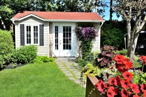 Ein helles Gartenhaus mit rotem Satteldach, weißer Sprossen-Doppeltür und einem Fenster mit schwarzen Fensterläden steht in einem grünen Garten. Ein Steinplattenweg führt durch den Rasen zur Tür, während im Vordergrund rote Petunien in einem Blumenkasten blühen und lila Clematis an der Fassade rankt.