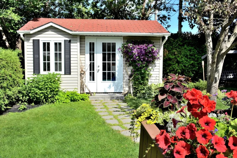 Ein helles Gartenhaus mit rotem Satteldach, weißer Sprossen-Doppeltür und einem Fenster mit schwarzen Fensterläden steht in einem grünen Garten. Ein Steinplattenweg führt durch den Rasen zur Tür, während im Vordergrund rote Petunien in einem Blumenkasten blühen und lila Clematis an der Fassade rankt.