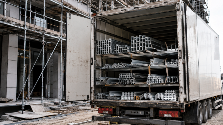Offener LKW mit gestapelten Aluminiumprofilen auf der Ladefläche neben einem Baugerüst an einer Hausfassade. Die Aluprofile werden für Bau- und Gerüstkonstruktionen auf der Baustelle angeliefert.