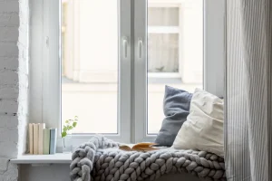 Modernes, weißes zweiflügeliges Kunststofffenster mit breiter Fensterbank, die als gemütliche Leseecke eingerichtet ist. Auf der Decke liegen Kissen, eine grob gestrickte Wolldecke und ein offenes Buch, links stehen dekorative Bücher.