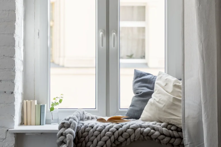 Modernes, weißes zweiflügeliges Kunststofffenster mit breiter Fensterbank, die als gemütliche Leseecke eingerichtet ist. Auf der Decke liegen Kissen, eine grob gestrickte Wolldecke und ein offenes Buch, links stehen dekorative Bücher.