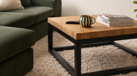 Massivholz-Couchtisch mit schwarzem Metallgestell in modernem Wohnzimmer mit dunkelgrüner Cord-Couch, hellem Langflorteppich, gestreifter Tasse und Magazinen.