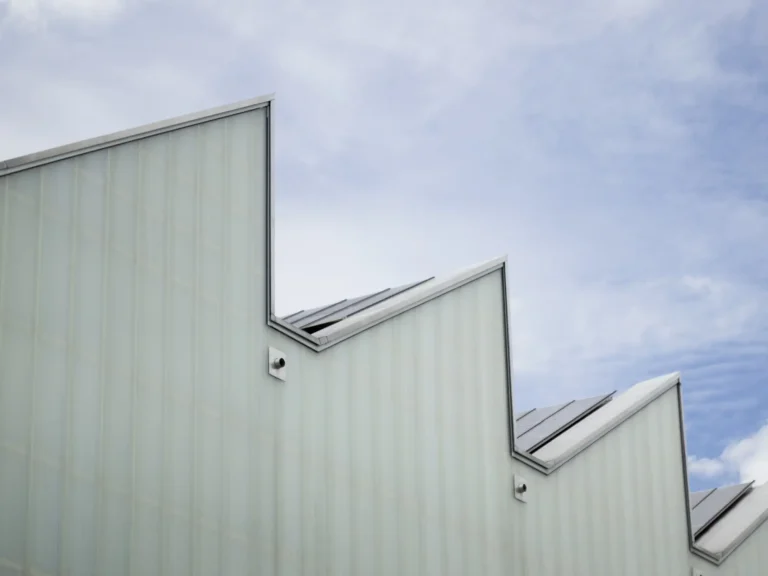 Moderne Industriehalle mit einem charakteristischen Sheddach. Die Fassade besteht aus hellen, vertikal strukturierten Profilbauglaselementen (U-Profilglas) vor einem leicht bewölkten, blauen Himmel.