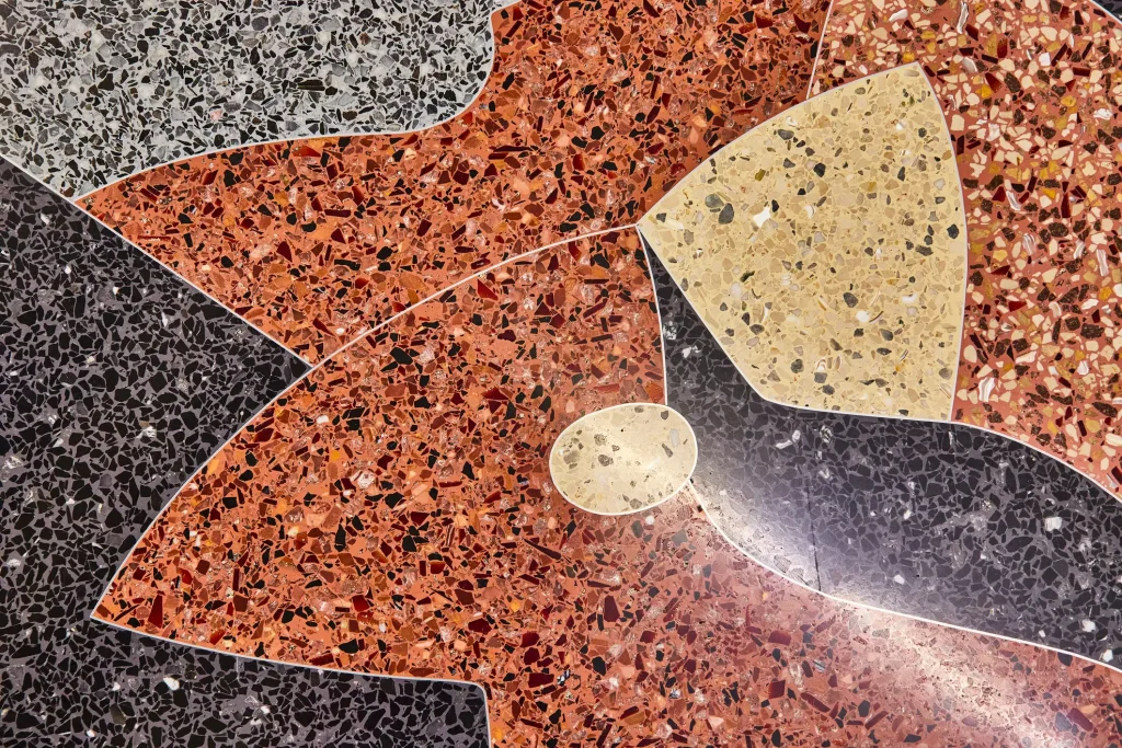 Nahaufnahme eines mehrfarbigen Terrazzobodens mit einem kunstvollen Mosaik-Muster. Kontrastierende, geschwungene Felder in Terrakotta-Rot, Dunkelgrau, Hellgrau und Beige zeigen die typisch gesprenkelte Struktur von mineralischen Einschlüssen.