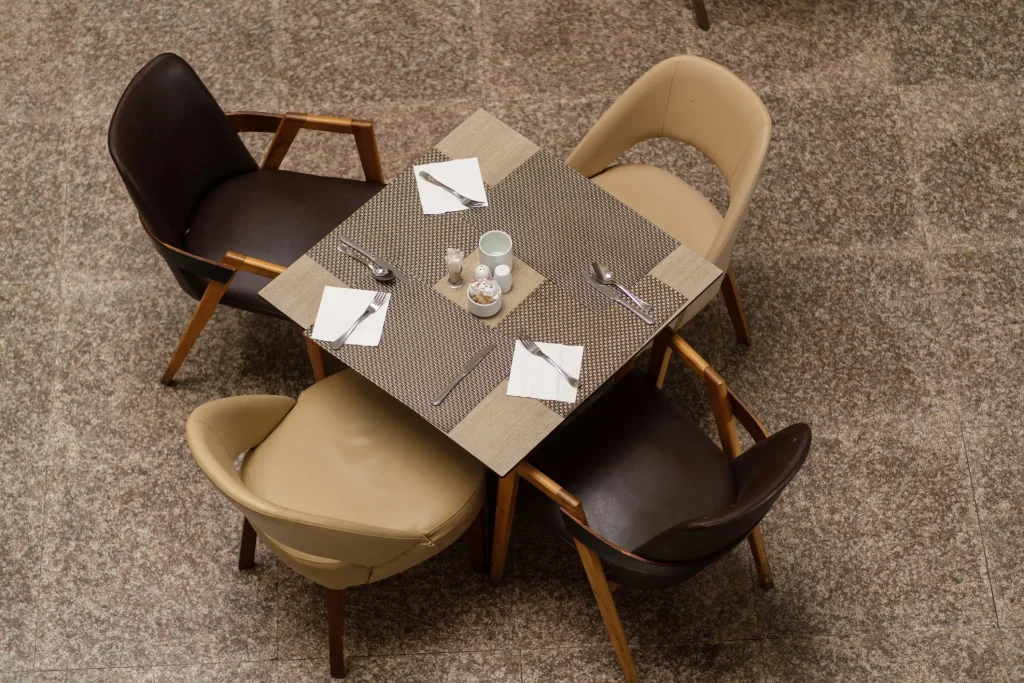 Draufsicht auf einen quadratischen, eingedeckten Restauranttisch mit vier Designerstühlen. Die gesamte Essgruppe steht auf einem glänzenden, rötlich-braun gesprenkelten Terrazzoboden in Granitoptik.