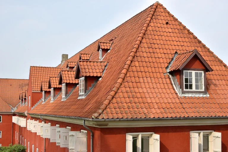 Historisches, rotes Gebäude mit einem großen, steilen Walmdach aus roten Tondachziegeln. In das ziegelgedeckte Dach sind mehrere kleine Schleppgauben mit weißen Fenstern integriert, und an der Fassade befinden sich geöffnete, helle Fensterläden.