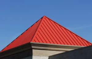 Modernes, pyramidenförmiges Zeltdach aus rotem Stehfalzblech auf einem Gebäude vor einem klaren, blauen Himmel.