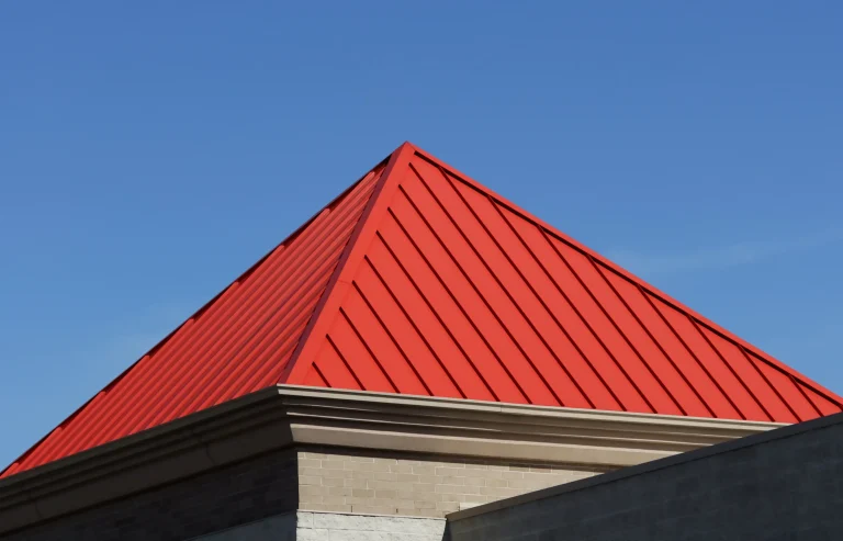Modernes, pyramidenförmiges Zeltdach aus rotem Stehfalzblech auf einem Gebäude vor einem klaren, blauen Himmel.