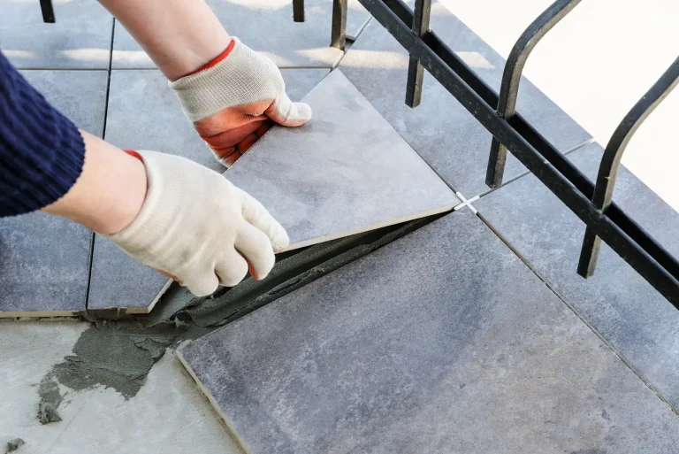 Nahaufnahme eines Fliesenlegers mit Arbeitshandschuhen, der eine quadratische, grau melierte Fliese präzise im Fliesenbett auf einem Balkon verlegt. Ein weißes Fliesenkreuz dient als Abstandshalter am Rand eines schwarzen Metallgeländers.