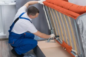 Ein Möbelmonteur in blauer Latzhose montiert den Klappmechanismus beziehungsweise das Bettkasten-Scharnier eines orangen Schlafsofas bei der Möbelmontage.