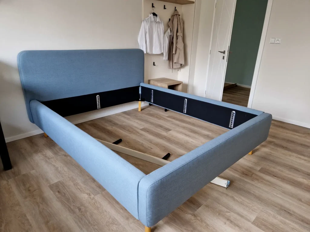 Montierter blauer Polsterbett-Rahmen mit gepolstertem Kopfteil ohne Lattenrost und Matratze auf einem hellen Holzboden im Schlafzimmer.
