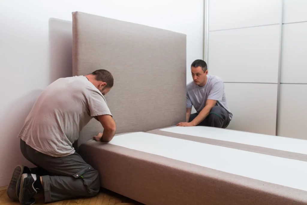 Zwei Möbelmonteure befestigen gemeinsam das gepolsterte Kopfteil an einem beigen Boxspringbett bei der Möbelmontage im Schlafzimmer.