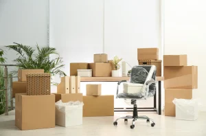 Gepackte Umzugskartons stapeln sich in einem hellen Büro während einer Büroauflösung. Im Zentrum steht ein in Stretchfolie verpackter Bürostuhl auf Rollen vor einem Schreibtisch, umgeben von Kisten, Ordnern und Verpackungsmaterial.