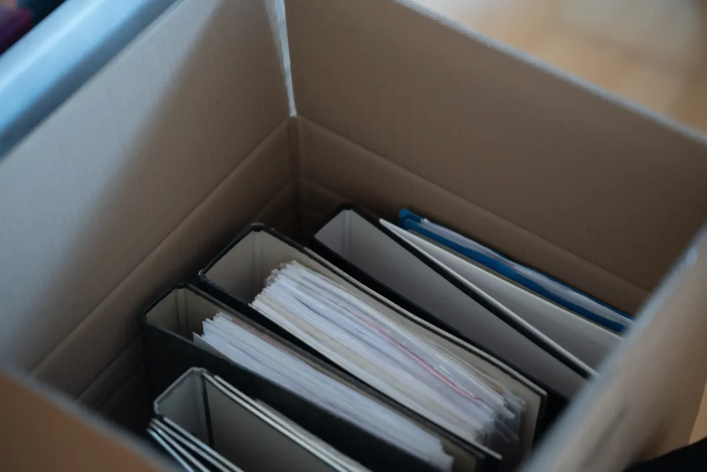 Blick von oben in einen geöffneten Umzugskarton, in dem mehrere schwarze Aktenordner mit Dokumenten und Papierstapeln nebeneinander einsortiert sind.