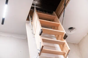 Blick von unten auf eine ausgeklappte, dreiteilige Holztreppe einer Dachbodentreppe (Bodentreppe), die in die geöffnete Deckenluke eines Dachbodens führt.