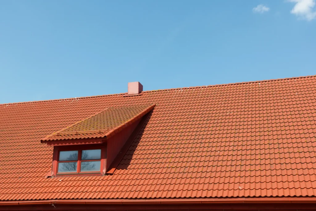 Außenansicht einer gleichmäßigen Dacheindeckung mit roten Dachpfannen auf einem Pfettendach. In die Dachfläche ist eine Schleppgaube mit einem kleinen Sprossenfenster integriert, darüber ragt ein kurzer, roter Schornstein vor einem klaren, blauen Himmel auf.