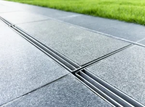 Moderne, dezent integrierte Entwässerungsrinne aus Edelstahl auf einer Garten-Terrasse mit grauen Natursteinplatten direkt neben einer Rasenfläche.