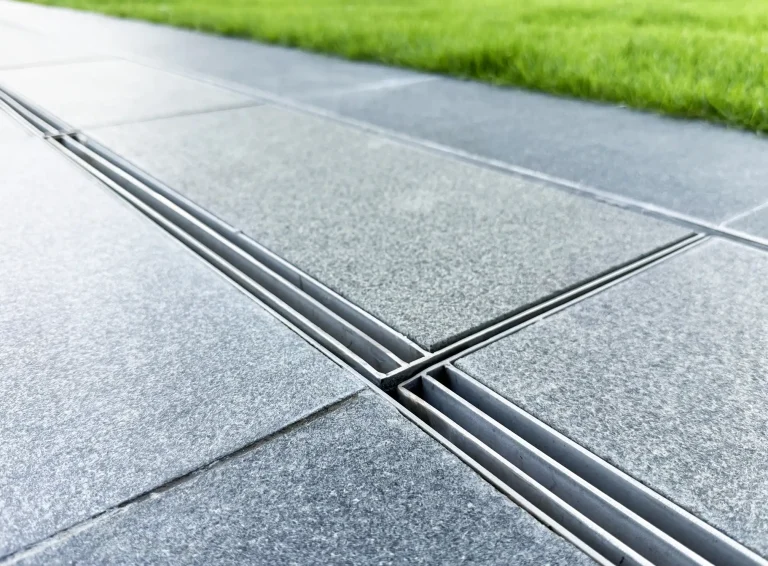 Moderne, dezent integrierte Entwässerungsrinne aus Edelstahl auf einer Garten-Terrasse mit grauen Natursteinplatten direkt neben einer Rasenfläche.